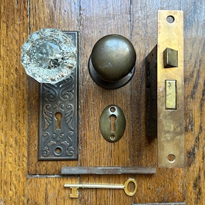 Op de afbeelding: Een verzameling antiek deurbeslag bestaande uit een heldere glazen knop op een rechthoekige donkere metalen achterplaat, een ronde messing knop, een messing insteekslot, een klein ovaal sleutelplaatje, een metalen stift en een messing sleutel.