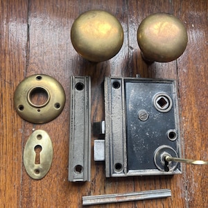 May include: An assortment of antique door hardware, including two brass doorknobs, a door lock, a strike plate, a keyhole cover, and a latch plate. The hardware is arranged on a wooden surface, showcasing its vintage design.