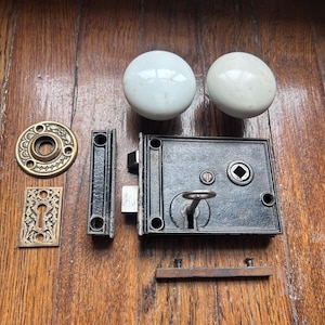 Puede incluir: Juego de herrajes de puerta antiguos que incluye dos pomos de cerámica blanca, una cerradura de metal negro y placas de latón. El conjunto está dispuesto sobre una superficie de madera, mostrando el diseño y los componentes vintage.