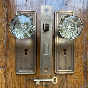 Pode incluir: Um conjunto de duas placas de porta em latão com puxadores de vidro transparente facetado, um mecanismo central de fechadura de embutir em latão com o texto Yale e uma chave. As placas retangulares possuem um buraco de fechadura e furos.
