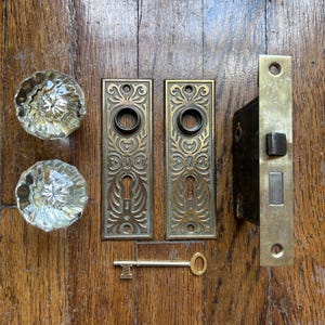 May include: A collection of antique door hardware, featuring two clear glass doorknobs, two decorative brass doorplates, a brass key, and a brass door latch. The items are displayed on a wooden surface.