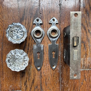 May include: Antique door hardware set including two clear glass doorknobs, two metal escutcheons, and a metal door latch. The hardware is set against a wooden background.