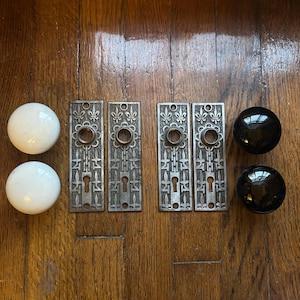 May include: Two white spherical door knobs, two black spherical door knobs, and four rectangular metal backplates featuring a raised fleur-de-lis pattern and a central keyhole opening, arranged in a row on a wooden surface.
