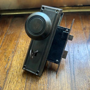 May include: A dark bronze-toned metal door knob set featuring a circular knob, a rectangular backplate with screw holes at the corners, and a mortise lock mechanism with a small skeleton key inserted into the keyhole on the front plate.