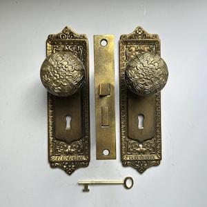 May include: A set of two brass door knobs with matching rectangular backplates featuring ornate embossed floral patterns and keyholes, a central rectangular latch mechanism, and a single brass skeleton key positioned below.
