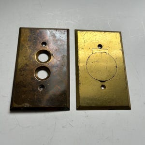 Pode incluir: Duas placas de parede retangulares de latão. A placa esquerda tem uma pátina escura e manchada com duas aberturas circulares e dois pequenos orifícios para parafusos. A placa direita tem um acabamento polido com uma tampa circular.