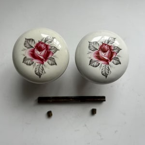 May include: Two round white ceramic door knobs featuring a painted red rose with grey leaves. The set includes a long dark metal spindle and two small metal set screws positioned below the knobs on a plain surface.