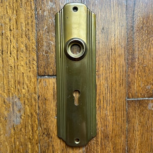 May include: A rectangular brass door plate featuring a central circular knob opening and a lower keyhole cutout. The plate has rounded top and bottom edges with vertical grooved detailing along the sides and two small screw holes for mounting.