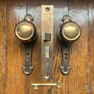 May include: A set of two brass door knobs with matching backplates, a central rectangular mortise lock mechanism, and a single brass skeleton key. The hardware features a polished metallic finish with visible screw holes for mounting purposes.