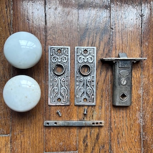 May include: Antique door hardware set, including two white porcelain doorknobs, two ornate metal backplates, a metal latch, and a metal strike plate. The hardware is arranged on a wooden surface.