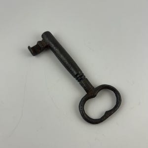 May include: An antique iron skeleton key with a dark, weathered finish. The key features a rounded, oval-shaped bow, a cylindrical shank with a small decorative collar near the base, and a distinct, notched bit at the end for a mechanical lock.