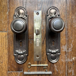 Pode incluir: Conjunto de ferragens de porta em bronze antigo, incluindo duas maçanetas, uma placa de fecho, uma chave e uma tranca. As maçanetas são redondas e montadas em placas retangulares com detalhes decorativos. A chave é dourada.