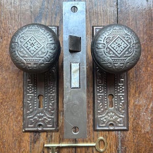 May include: Antique door hardware set featuring two ornate doorknobs with decorative plates, a central latch, and a brass key. The metal components have a weathered, aged appearance. The door hardware is mounted on a wooden door.