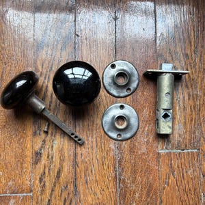 Puede incluir: Un surtido de herrajes de puertas antiguos, que incluyen dos pomos de puerta de vidrio negro, dos placas traseras circulares de metal y un pestillo de puerta de metal. El hardware está dispuesto sobre una superficie de madera.