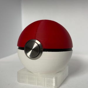 May include: A red and white sphere with a silver button, resembling a Pokeball. The sphere is set on a clear, rectangular base. The top half is red, the bottom half is white, and a black band separates the two colors.