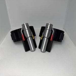 May include: A pair of black wrist cuffs featuring silver-toned cylindrical attachments. Each attachment is accented with a small red rod on one side and a yellow rod on the other, mounted on a black rectangular base secured to the wide black wristband.