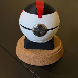 May include: A white and black sphere with a red stripe on top and a silver button in the center. The sphere sits on a black square base, which is on a cork base. The item is likely a decorative object or a unique gadget.