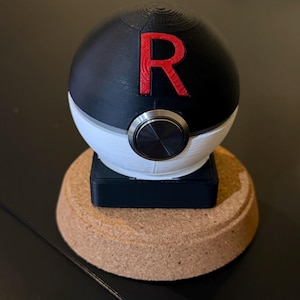 Puede incluir: Una esfera negra y blanca con una "R" roja en la parte superior, que se asemeja a una Poké Ball. Tiene un botón plateado y se asienta sobre una base negra encima de un posavasos de corcho. El diseño está inspirado en Pokémon.