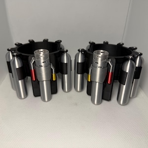 May include: A pair of black circular wrist cuffs featuring multiple silver-colored cylindrical canisters attached to the exterior. Each cuff includes a central silver-colored nozzle component with small red and yellow accents on the sides.