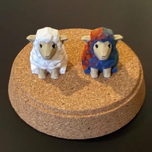 May include: Two carved wooden sheep figurines. One sheep is white, the other has a blue, red, and orange pattern. Both have tan faces and stand on a round, cork base. The background is black.