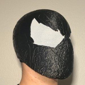 May include: A black full-head mask featuring a textured, organic surface pattern and large, white mesh eye lenses. The mask covers the entire head and face, with a distinct jagged, vein-like relief texture across the exterior surface.