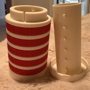 May include: A white and red puzzle cylinder with rotating bands of letters, next to a white cylindrical container. The puzzle has red bands with white letters. The cylindrical container has a circular base.