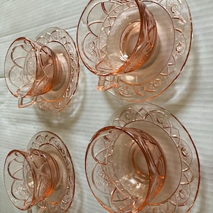 May include: A set of four pink depression glass teacups and matching saucers. Each piece features a molded floral pattern with a scalloped edge design. The translucent pink glass has a glossy finish and a consistent geometric motif throughout.