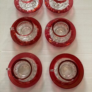 Puede incluir: Un juego de seis tazas de té y platillos a juego de cristal transparente, cada uno con un borde rojo vibrante y un patrón texturizado de pequeñas hendiduras circulares en el exterior de las tazas. Las tazas tienen pequeñas asas.