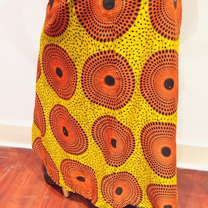 May include: A vibrant yellow dress with a repeating pattern of orange and black circular designs. The dress has a flowing silhouette and is made of a lightweight fabric. The design is reminiscent of tribal art.