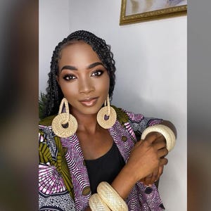 Puede incluir: Una mujer con una chaqueta estampada en morado y verde con grandes pendientes de paja trenzada y pulseras de paja trenzada a juego.