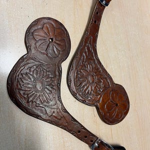 May include: Two brown leather spurs with floral designs. The spurs feature embossed sunflowers and intricate leaf patterns. Each spur has a strap with a silver buckle for secure attachment to riding boots. The leather has a rich, warm tone.