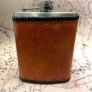 May include: A hip flask with a brown leather casing and a silver-coloured cap. The leather is stitched with black thread. The flask has a hinged closure and is designed to hold liquid.