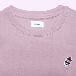 Puede incluir: Una sudadera de color malva con cuello redondo y un pequeño parche bordado de helado en el pecho. La etiqueta dice "jak_squat". Esta prenda informal es adecuada para el uso diario.