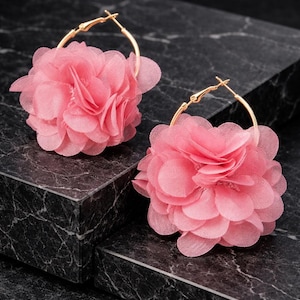 Può includere: Un paio di orecchini a cerchio con fiori rosa. Ogni orecchino presenta un cerchio color oro con un grande fiore di tessuto rosa a strati. Gli orecchini sono esposti su una superficie scura e marmorata.