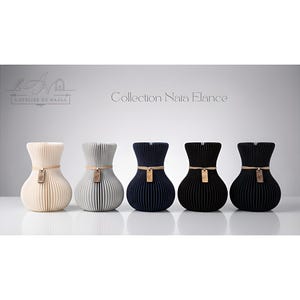Peut inclure: Une collection de cinq vases décoratifs de couleurs différentes. Les vases sont beiges, gris, bleu marine et noirs. Chaque vase a une étiquette et une corde autour du col. Le texte "Collection Nara Elance" est au-dessus des vases.