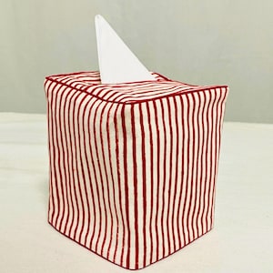 Puede incluir: Una funda cuadrada para caja de pañuelos con un patrón de rayas rojas y blancas. La funda es de tela y tiene un pañuelo blanco que sobresale de la parte superior.