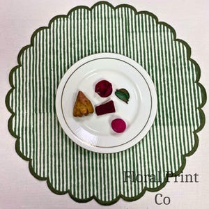 Può includere: Una tovaglietta rotonda con bordo smerlato caratterizzata da un motivo a righe verticali verdi e bianche. Al centro della tovaglietta si trova un piatto bianco con un sottile bordo dorato, contenente quattro piccoli pezzi di pot-pourri secco rosso, marrone e verde.