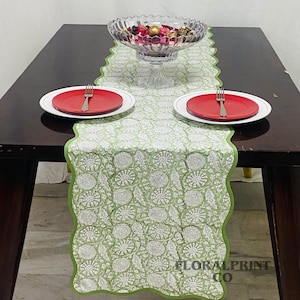 Peut inclure: Une table dressée avec un chemin de table à motifs floraux verts et blancs, bordé de festons. Deux assiettes rouges avec des fourchettes sont de chaque côté du chemin de table. Un bol en verre rempli d'objets décoratifs est au centre.