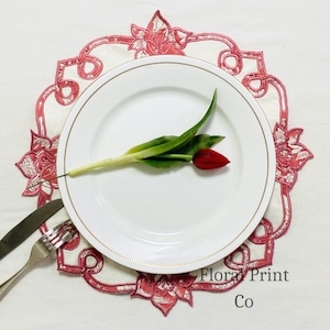 Può includere: Un piatto piano bianco con un sottile bordo dorato poggia su una tovaglietta rotonda bianca con un bordo ricamato a motivi floreali rossi. Un tulipano rosso con foglie verdi è adagiato sul piatto. Sono visibili un coltello e una forchetta d'argento. Il testo recita Floral Print Co.