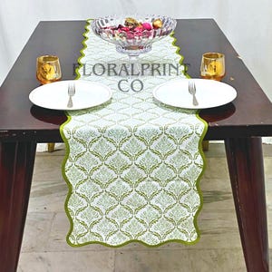 Peut inclure: Une table à manger dressée pour deux, avec un chemin de table à motifs blancs et verts avec un bord festonné. Comprend des assiettes blanches, des couverts, des bougeoirs dorés et un bol en verre rempli d'objets décoratifs. Le texte "FLORALPRINT CO" est visible.
