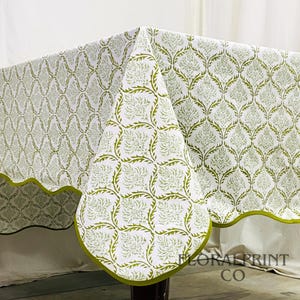 May include: A white tablecloth with a green floral pattern. The tablecloth has a scalloped edge with a green trim. The pattern is a repeating design of stylized leaves and flowers. The tablecloth is draped over a table.