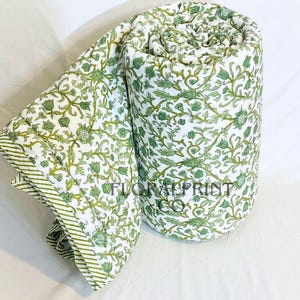 May include: Rolled-up white quilt with a green floral pattern. The quilt features a repeating design of green leaves and flowers. The edge of the quilt has a green and white striped border. The text "FLORAL PRINT" is visible on the quilt.