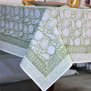 Puede incluir: Un mantel floral verde y blanco con un patrón detallado. El mantel presenta un diseño repetitivo de flores y hojas, con un borde decorativo. La tela está doblada, mostrando los intrincados detalles del patrón.