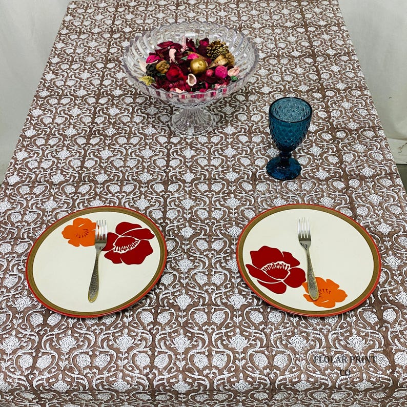 Rectangular Mud Brown cotton tablecloth with detailed floral hand block print and matching piping border spread across a farmhouse dining table. The handmade reusable table linen showcases authentic artisan craftsmanship and soft breathable cotton fabric, making it ideal for wedding table décor, seasonal gatherings, dinner parties, family meals, kitchen styling, and bohemian-inspired home decoration.