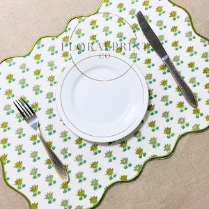 May include: A white placemat with a scalloped edge and a green border, adorned with a repeating pattern of green and yellow floral designs. A white plate with a gold rim, a fork, and a knife are on the placemat. The text "FLORAL PRINT CO" is visible.
