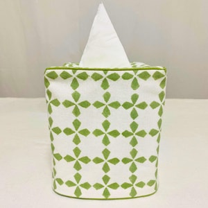 Puede incluir: Una funda de caja de pañuelos de tela con fondo blanco y un estampado floral verde repetido. El borde superior y el ribete son verdes. Los pañuelos blancos son visibles en la parte superior.