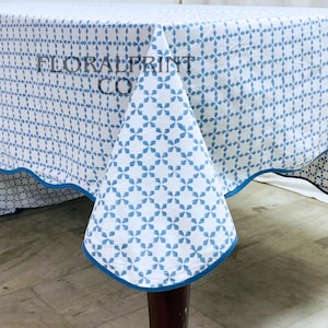 May include: White tablecloth with a blue geometric pattern and a blue trim. The tablecloth has the text "FLORALPRINT CO" printed on it. The tablecloth is draped over a wooden table.