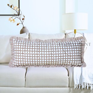 May include: A rectangular decorative pillow with a white background and a brown floral pattern. The pillow has a ruffled border in a striped brown fabric. The pillow is placed on a white sofa.