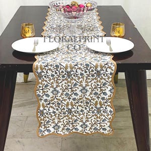 May include: A dining table set with a floral print table runner, white plates, and gold candle holders. A clear glass bowl filled with decorative items sits in the center. The table runner has a white background with blue and gold floral patterns.