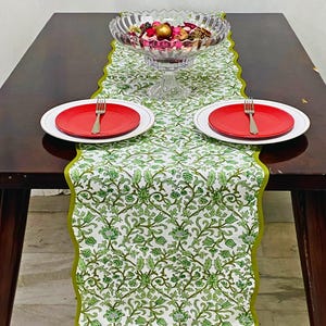 Peut inclure: Une table de salle à manger avec un chemin de table à imprimé floral vert et blanc. Deux assiettes rouges avec des fourchettes sont placées de chaque côté du chemin de table. Un bol en verre transparent rempli d'objets décoratifs est au centre.